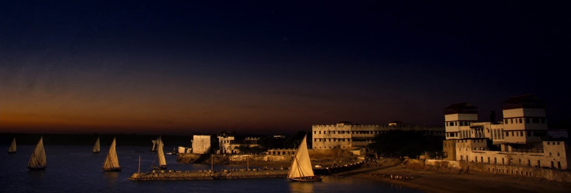 Lamu waterfront at dusk, dhows sailing past the historic fort and Swahili coast architecture